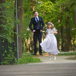 Foto Kubiczek - Chłopiec w garniturze i dziewczynka w białej sukience, oboje w podskoku na drewnianym mostku w Sosnowcu, otoczeni zielenią drzew.