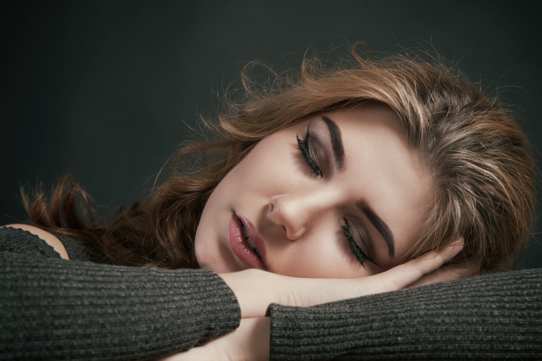 Portret śpiącej kobiety z delikatnym makijażem oczu, opierającej głowę na skrzyżowanych ramionach w szarym swetrze na ciemnym tle.