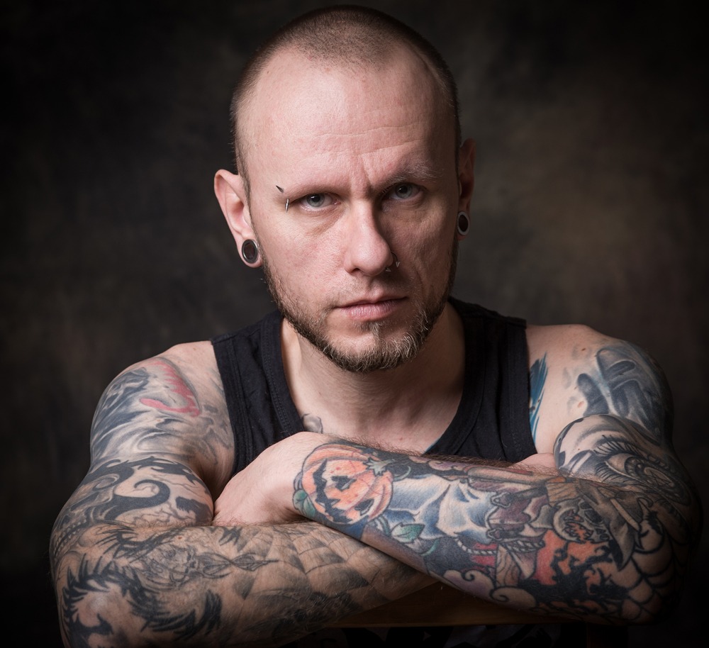 Portret mężczyzny z tatuażami na ramionach, kolczyk w brwi i nosie, czarny podkoszulek, ciemne tło.