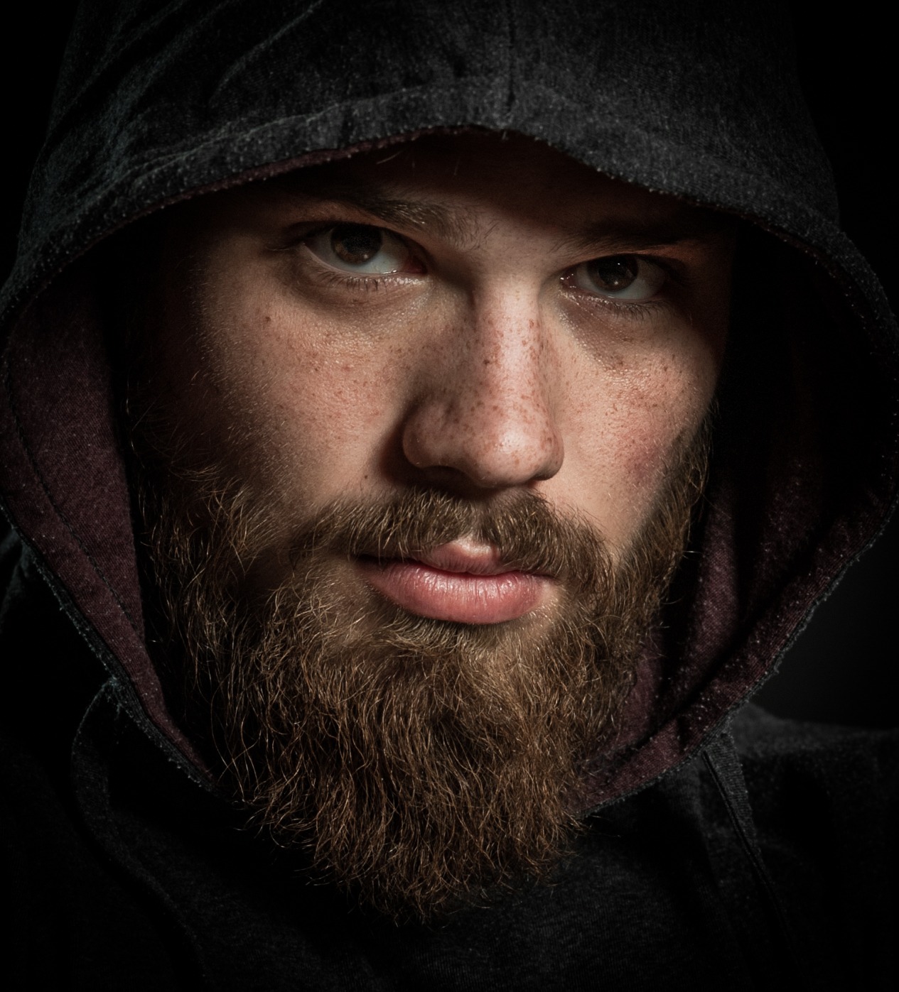Portret mężczyzny z brodą i piegami, w ciemnej bluzie z kapturem, oświetlony z góry