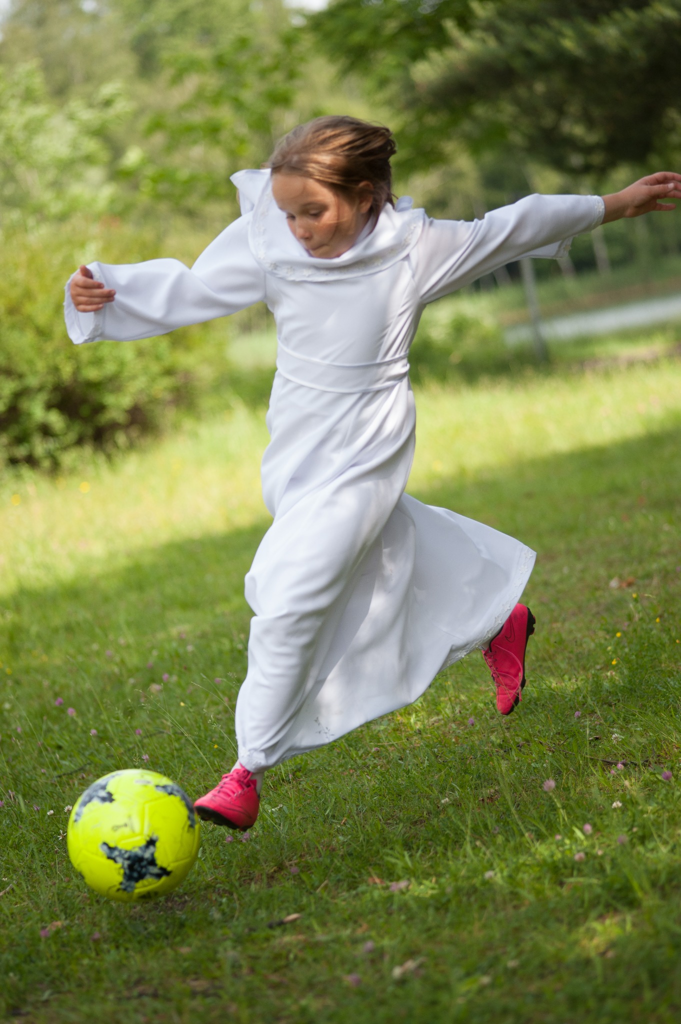 Dziewczynka w białej albie i różowych butach kopie żółto-czarną piłkę na zielonej trawie, rozłożone ramiona dla równowagi.
