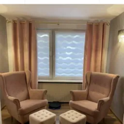 Pokój dzienny z dwoma różowymi fotelami uszakami, podnóżkami i oknem z dekoracyjną roletą dzień noc w faliste wzory, zasłoniętym różowymi zasłonami oraz z widocznym logo firmy na górze zdjęcia.