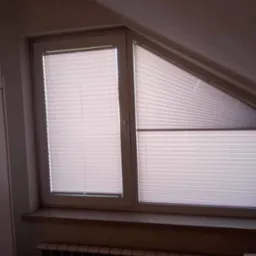 Dwa okna na poddaszu, jedno prostokątne, drugie trapezowe, osłonięte białymi, plisowanymi osłonami okiennymi, częściowo zasłoniętymi.