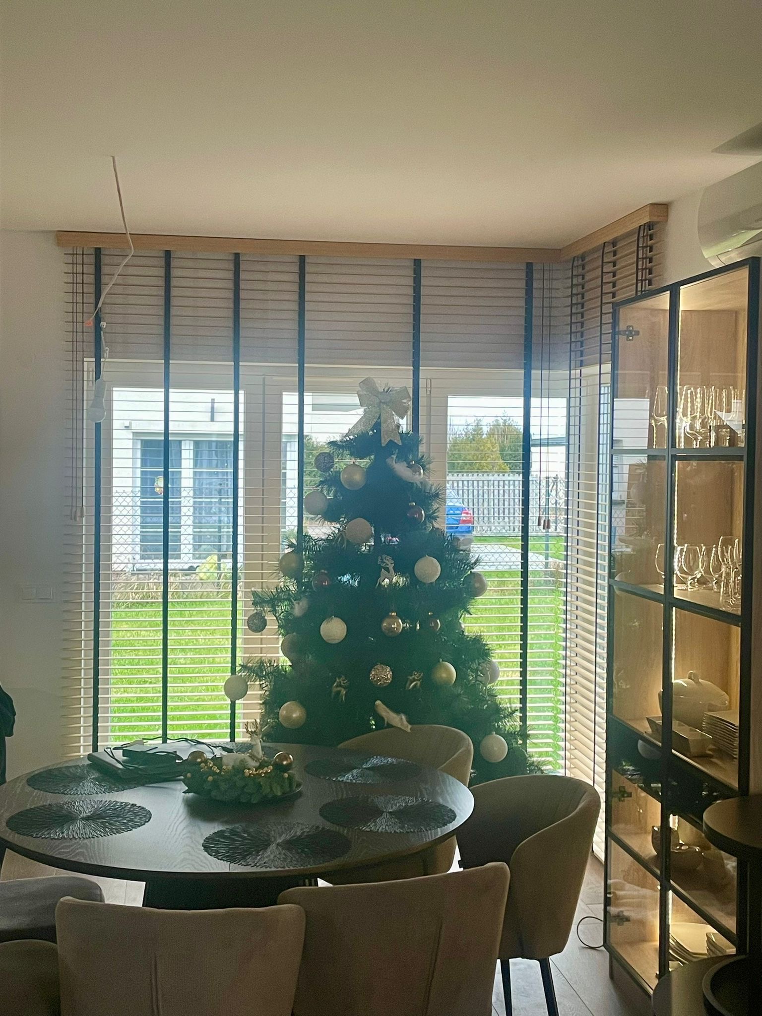 Jadalnia z okrągłym stołem i choinką świąteczną, widok przez okno z poziomymi żaluzjami drewnianymi w odcieniach beżu.