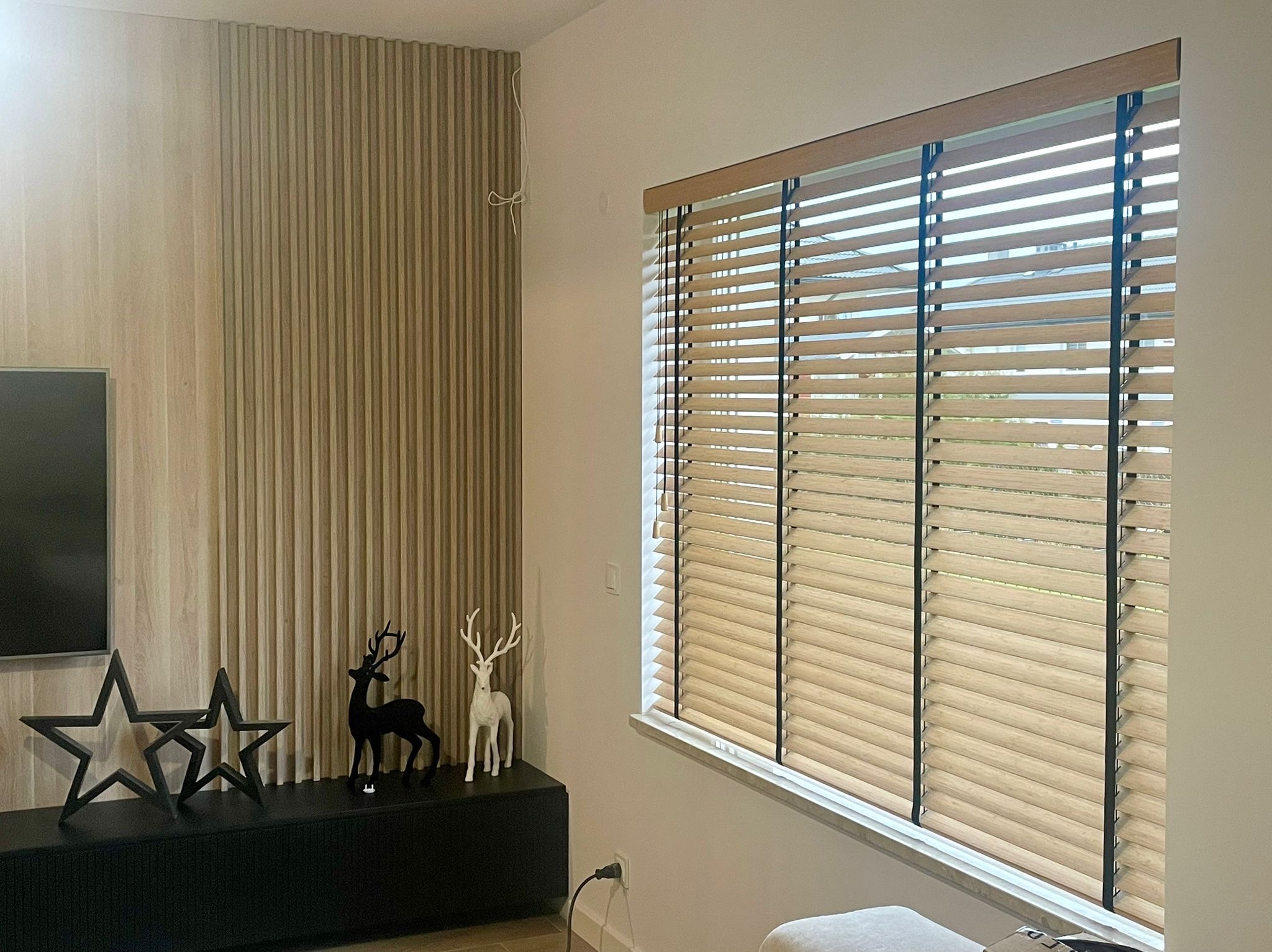 Jasne drewniane żaluzje poziome z czarnymi sznurkami zamontowane w oknie, widoczne elementy dekoracyjne: gwiazdy i figurki jeleni na szafce pod telewizorem.