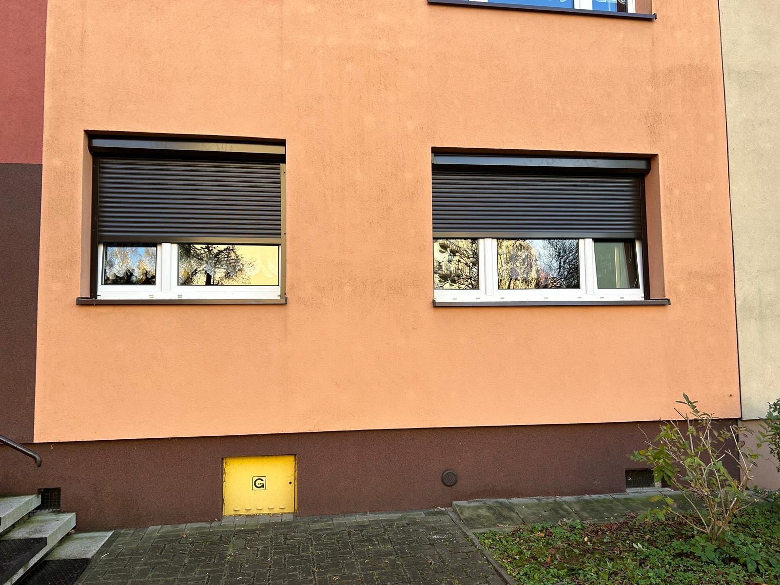 Dwa okna z opuszczonymi, ciemnymi przesłonami zewnętrznymi na pomarańczowej fasadzie budynku; w szybach okien odbijają się drzewa.