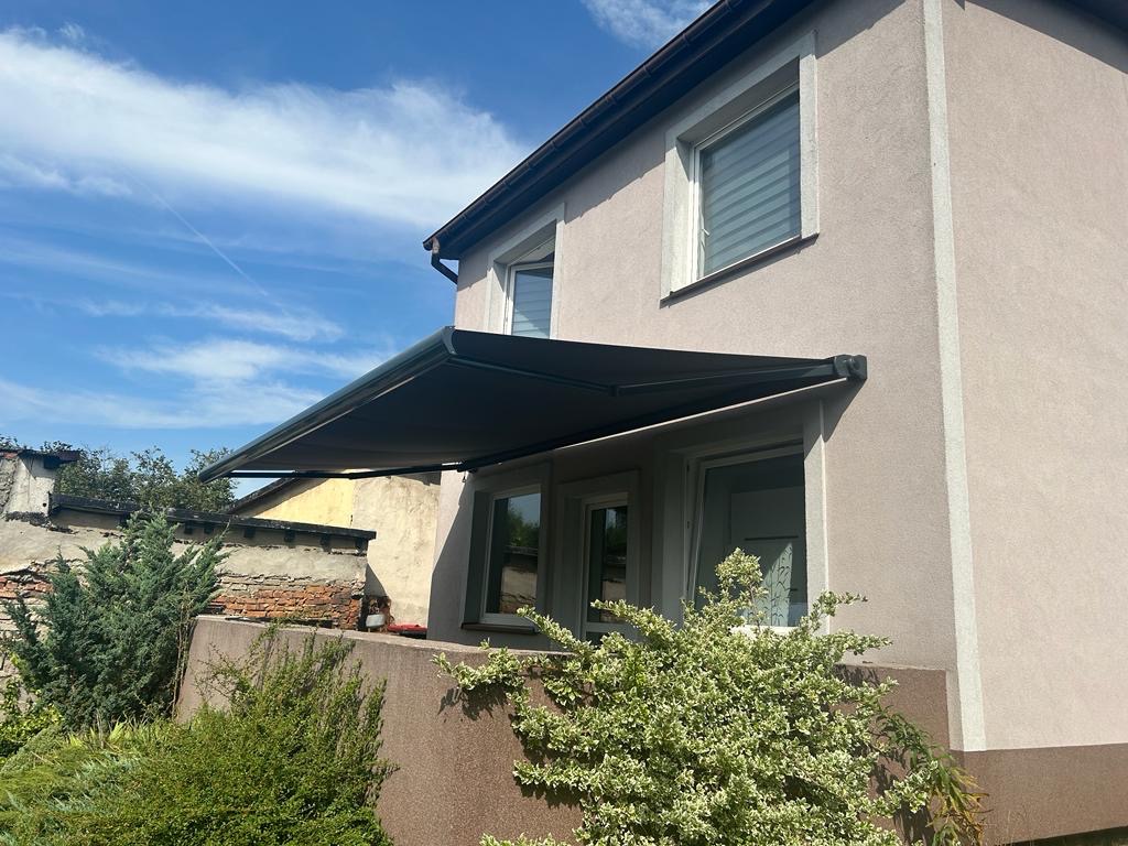 Ciemnoszara markiza tarasowa zamontowana nad balkonem domu jednorodzinnego z widocznymi żaluzjami w oknach na piętrze, błękitne niebo w tle.