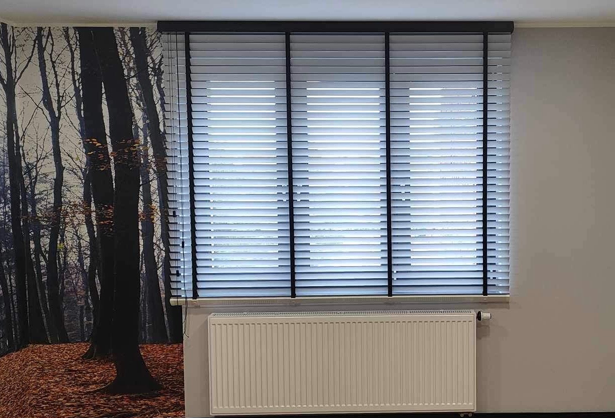 Zamknięte, poziome żaluzje aluminiowe w kolorze jasnym, z czarnymi sznurkami, zamontowane w oknie. Na ścianie widoczna fototapeta z motywem lasu.