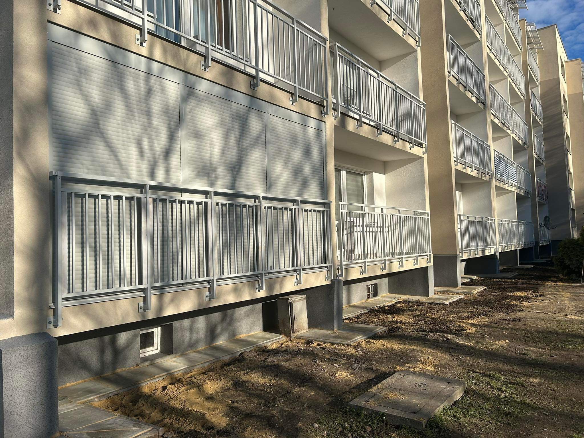 Fasada budynku wielorodzinnego z balkonami, na których zamontowano szare osłony okienne, widoczne z poziomu gruntu.