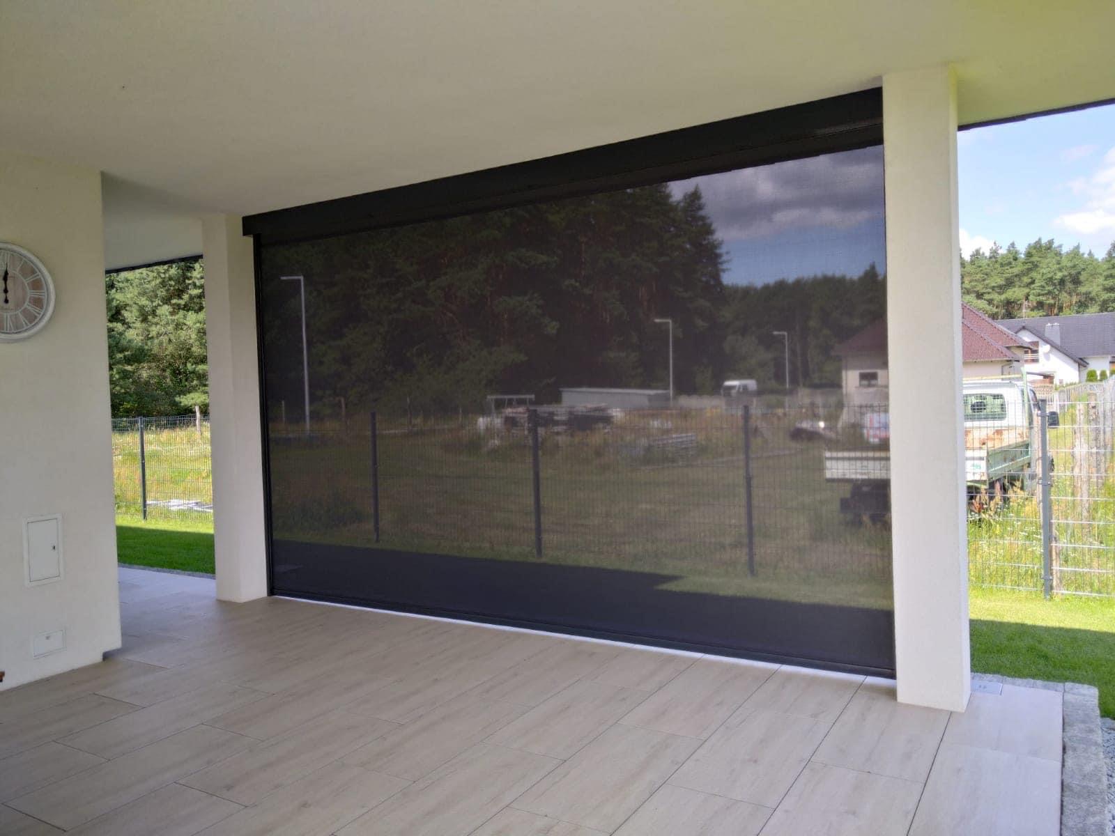 Zewnętrzna, zaciemniająca przesłona tarasowa w kolorze antracytowym, widok z zewnątrz na zalesiony teren i zabudowania.