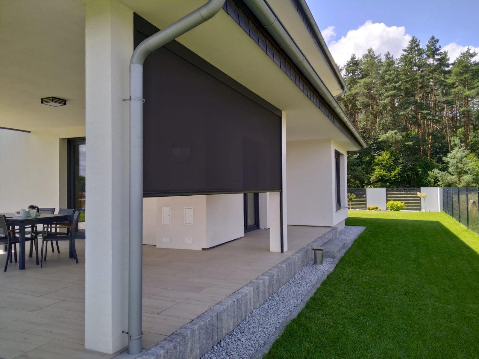 Ciemna, zwijana osłona przeciwsłoneczna na tarasie nowoczesnego domu z widokiem na zadbany trawnik i las w tle.