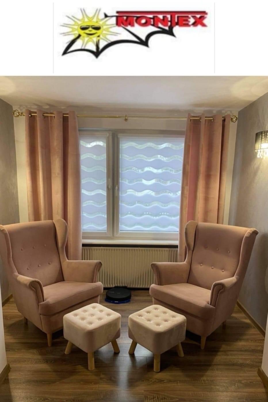 Pokój dzienny z dwoma różowymi fotelami uszakami, podnóżkami i oknem z dekoracyjną roletą dzień noc w faliste wzory, zasłoniętym różowymi zasłonami oraz z widocznym logo firmy na górze zdjęcia.