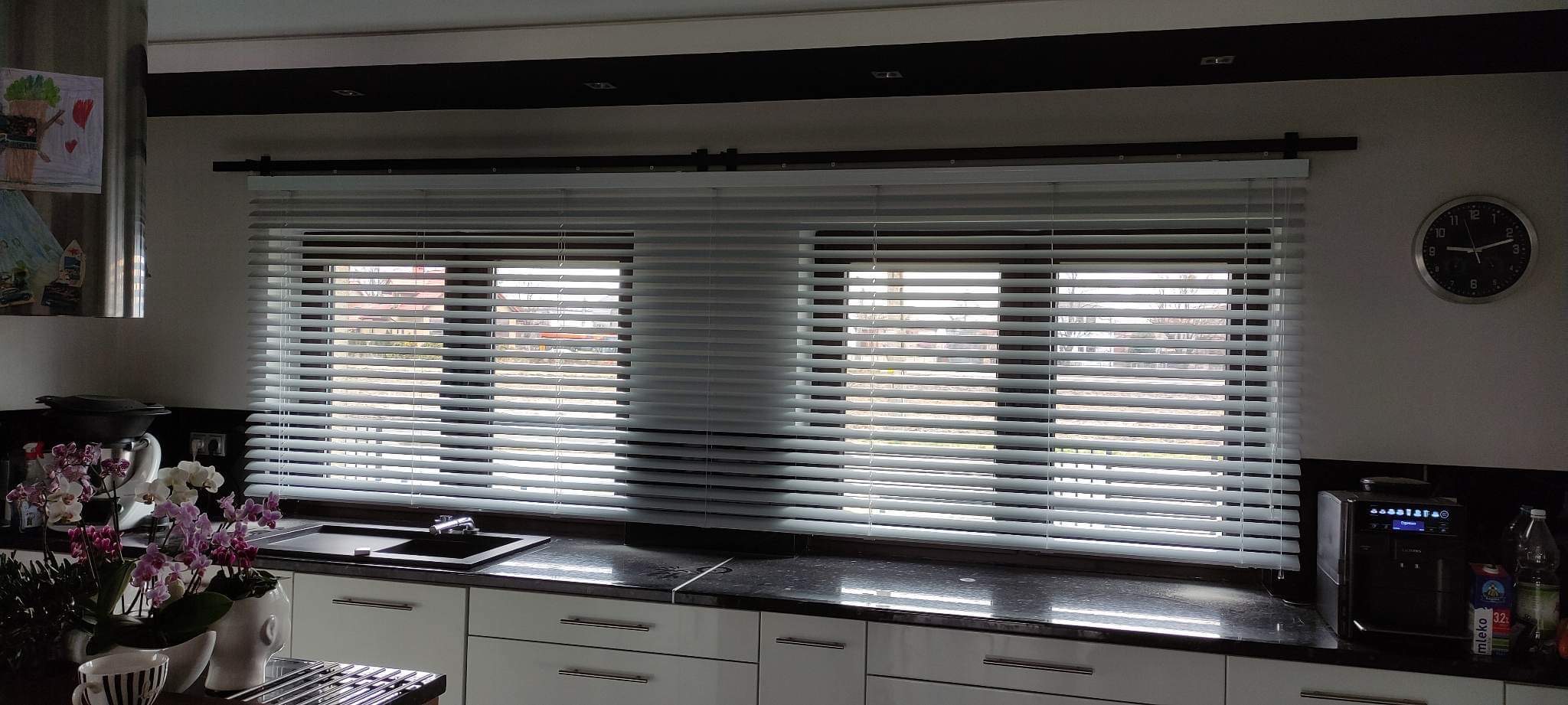 Kuchnia z dwoma oknami zasłoniętymi białymi żaluzjami aluminiowymi, na blacie kwiaty w doniczkach i ekspres do kawy.