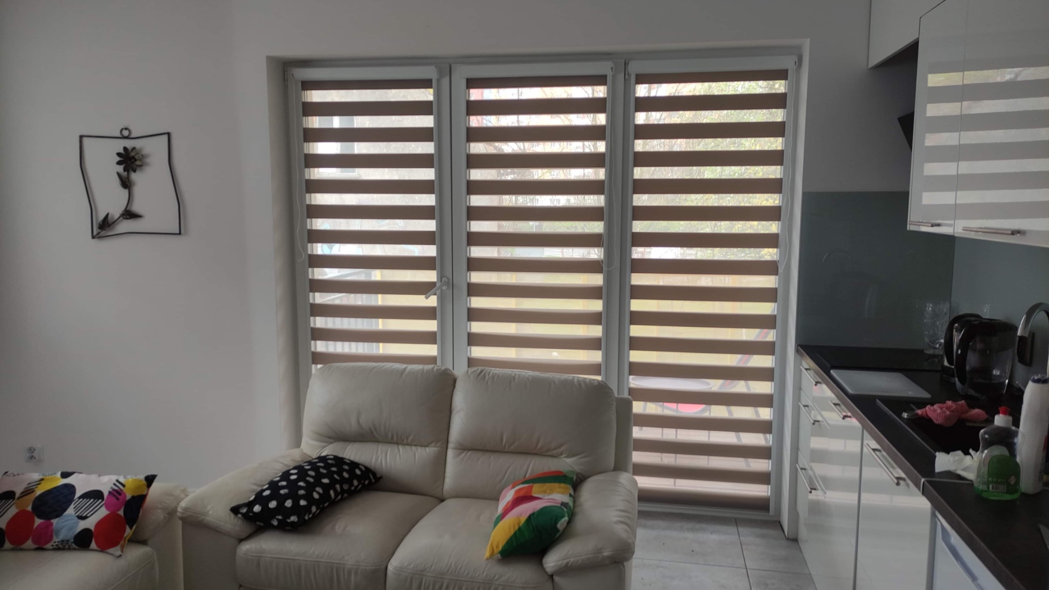 Jasne wnętrze w Zabrzu z trzema oknami przesłoniętymi roletami dzień noc w odcieniach beżu, widok z salonu na kuchnię.