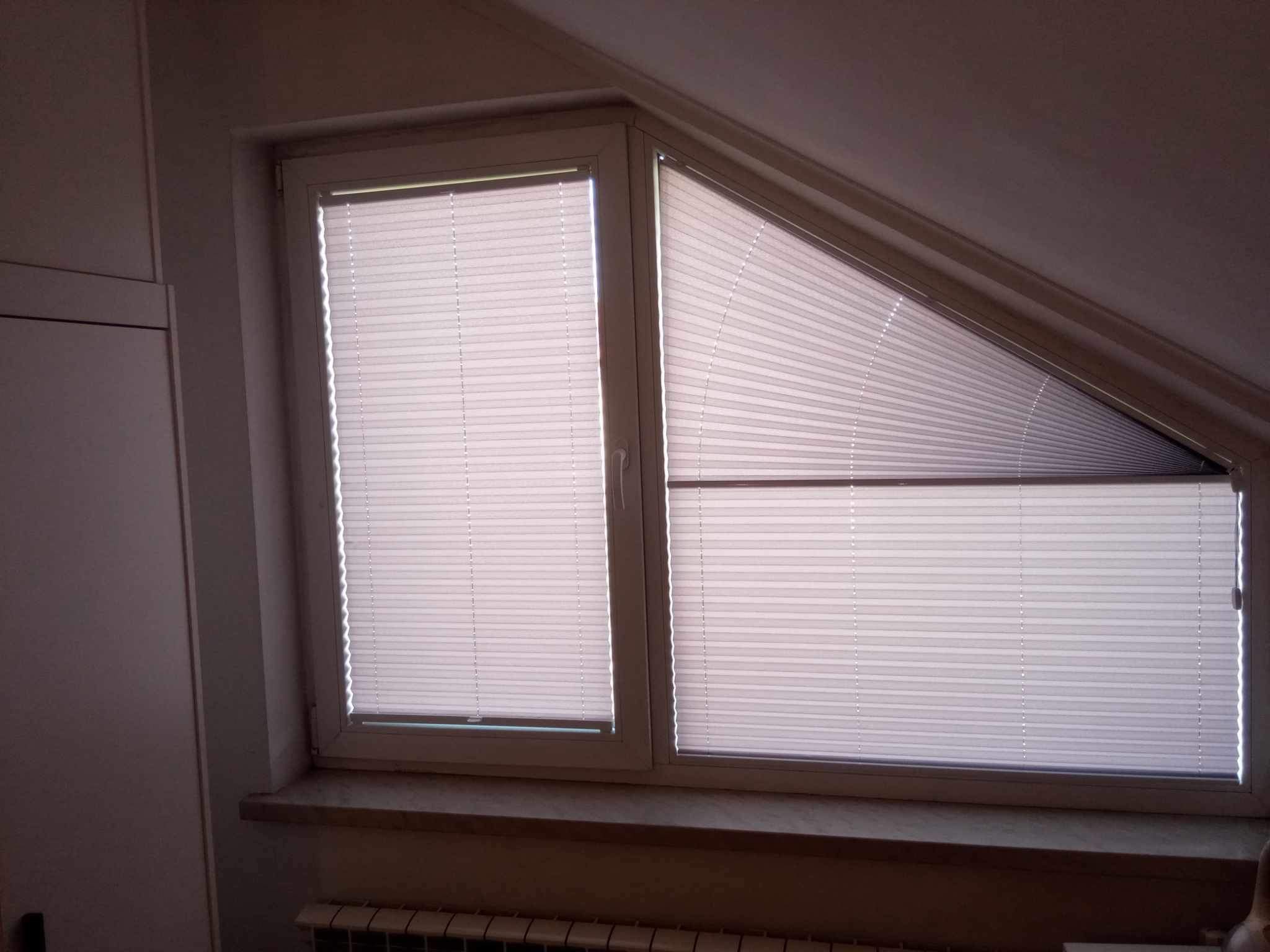 Dwa okna na poddaszu, jedno prostokątne, drugie trapezowe, osłonięte białymi, plisowanymi osłonami okiennymi, częściowo zasłoniętymi.