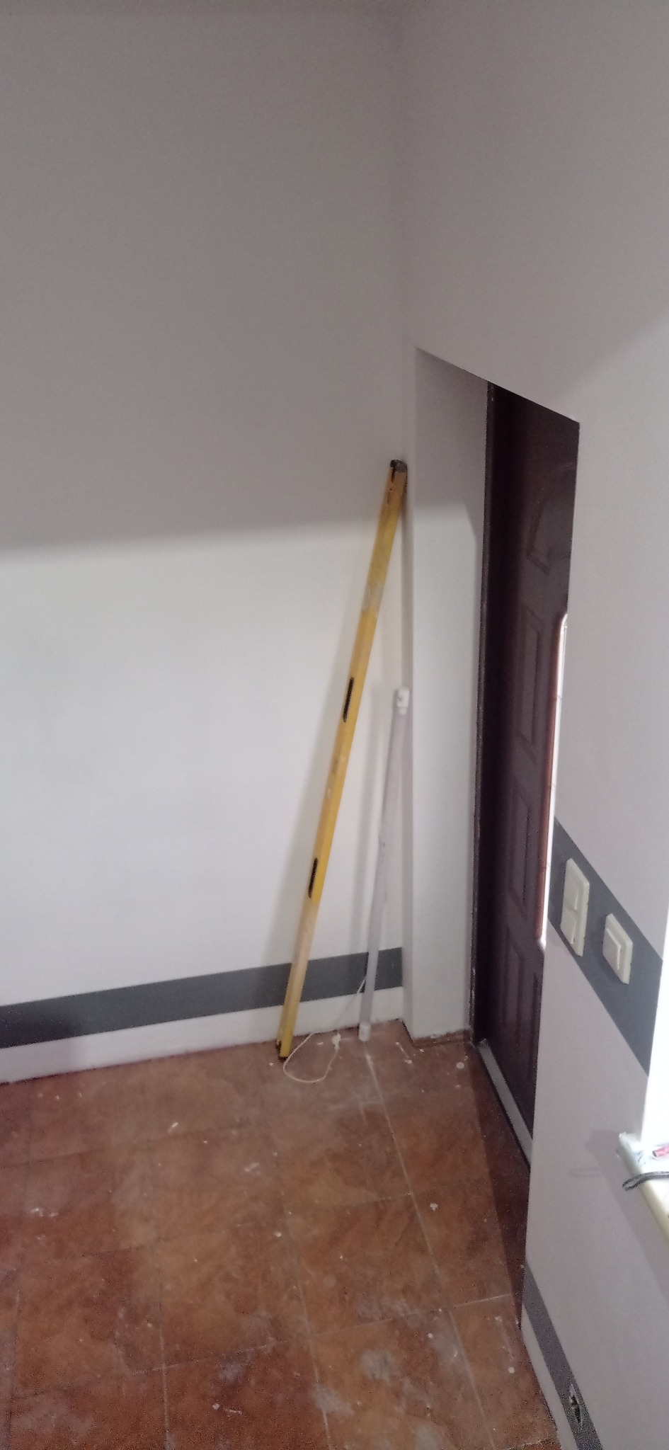 Wnętrze pokoju w trakcie remontu: poziomica oparta o ścianę, widoczne ślady po pracach na podłodze z terakoty i przy listwie przypodłogowej, częściowo widoczne drzwi.