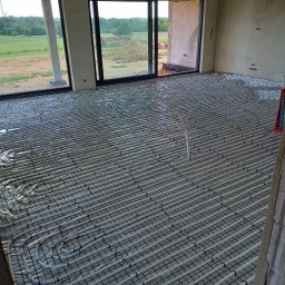 Instal-team - Rozłożona instalacja ogrzewania podłogowego na siatce w surowym wnętrzu z dużymi oknami wychodzącymi na zielony krajobraz.