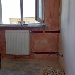 Instal-team - Montaż nowego grzejnika panelowego w bloku, widoczne czerwone rury instalacji centralnego ogrzewania prowadzone w ścianie, ściana w trakcie prac wykończeniowych, odkryte przewody elektryczne obok.