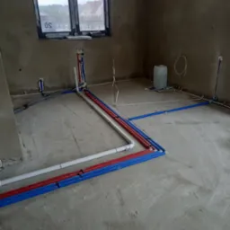 Surowy stan posadzki z rozłożonymi rurami instalacji wodnej i grzewczej w izolacji koloru niebieskiego i czerwonego, widoczne przyłącza do ściany i okno w tle.