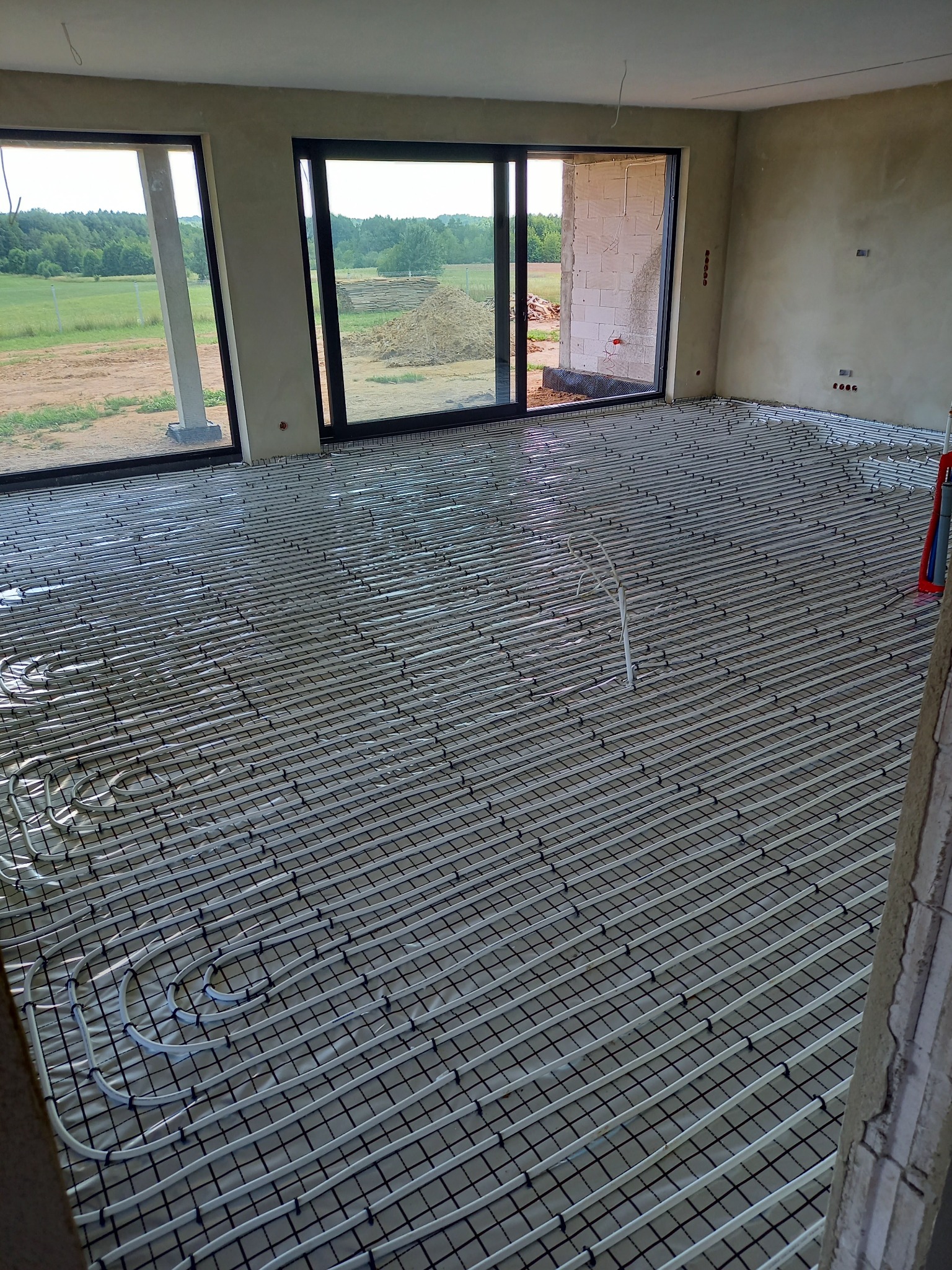 Rozłożona instalacja ogrzewania podłogowego na siatce w surowym wnętrzu z dużymi oknami wychodzącymi na zielony krajobraz.