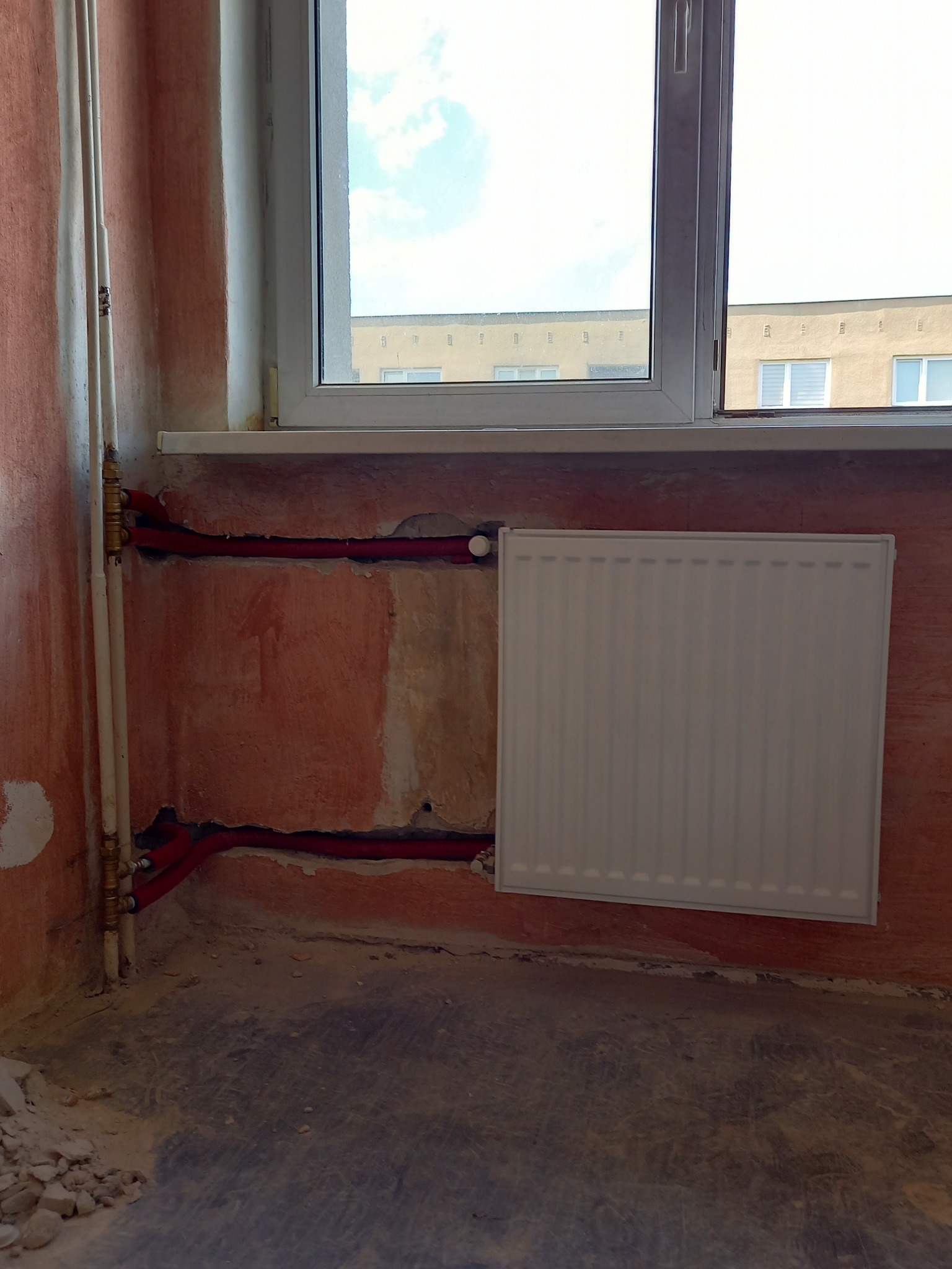 Świeżo zamontowany biały grzejnik panelowy w trakcie przeróbki instalacji c.o. w bloku, widoczne czerwone rury instalacyjne w ścianie i białe rury pionowe, ściany częściowo otynkowane, okno...