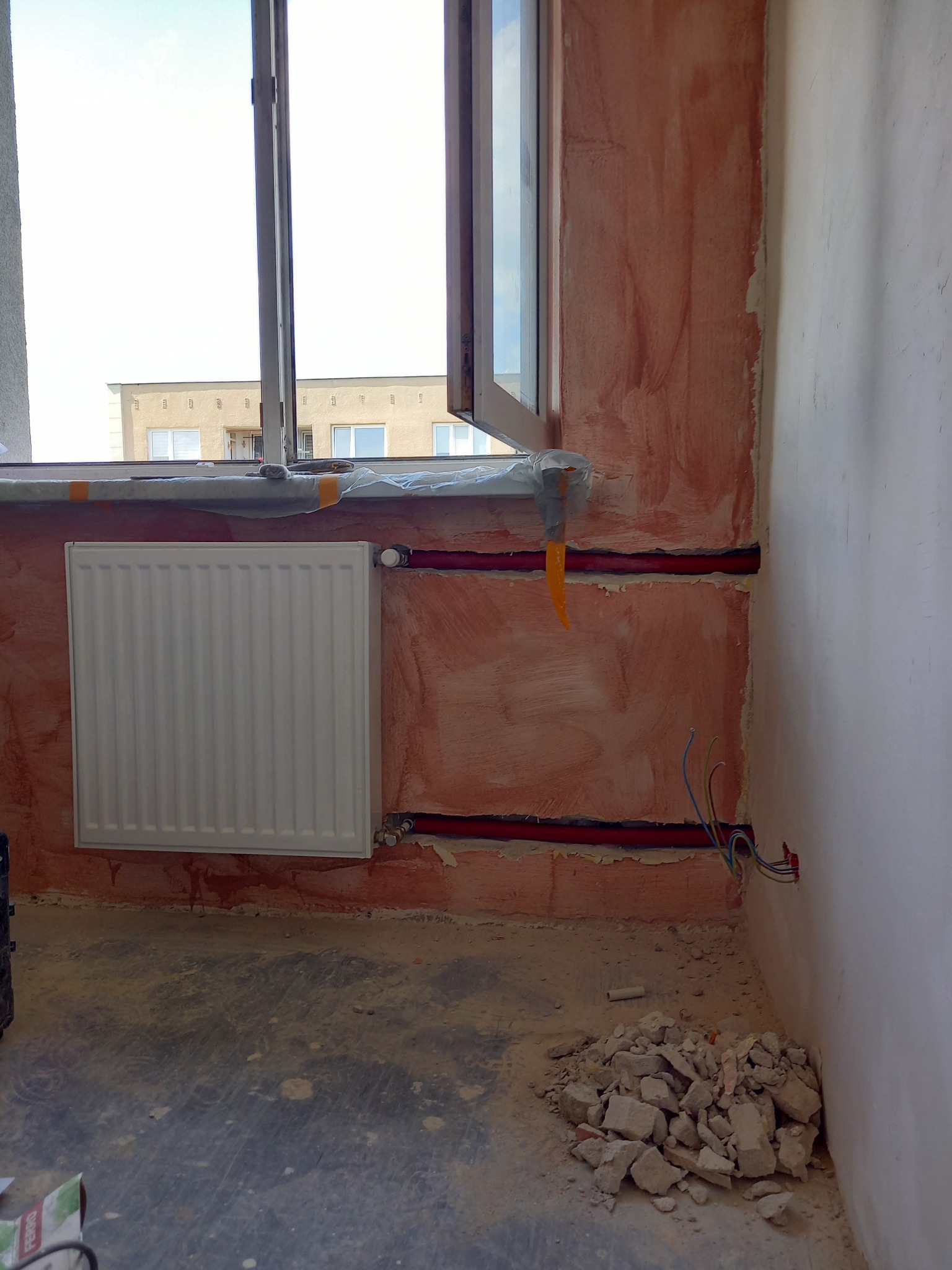 Montaż nowego grzejnika panelowego w bloku, widoczne czerwone rury instalacji centralnego ogrzewania prowadzone w ścianie, ściana w trakcie prac wykończeniowych, odkryte przewody elektryczne obok.