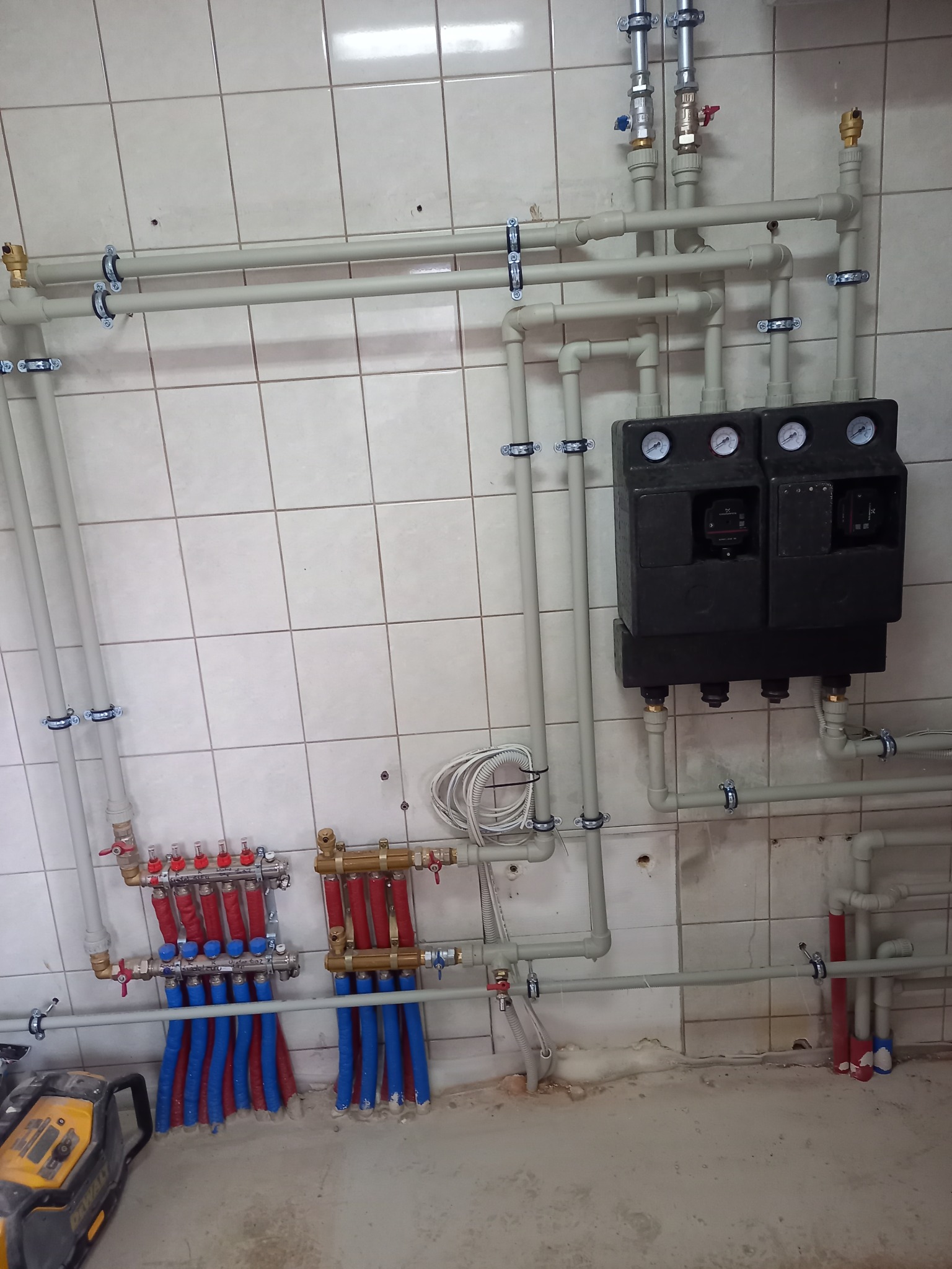 Rozdzielacz ogrzewania podłogowego z pompami obiegowymi zamontowany na ścianie z płytek, widoczne rury doprowadzające i odprowadzające wodę oraz narzędzie Dewalt na podłodze.
