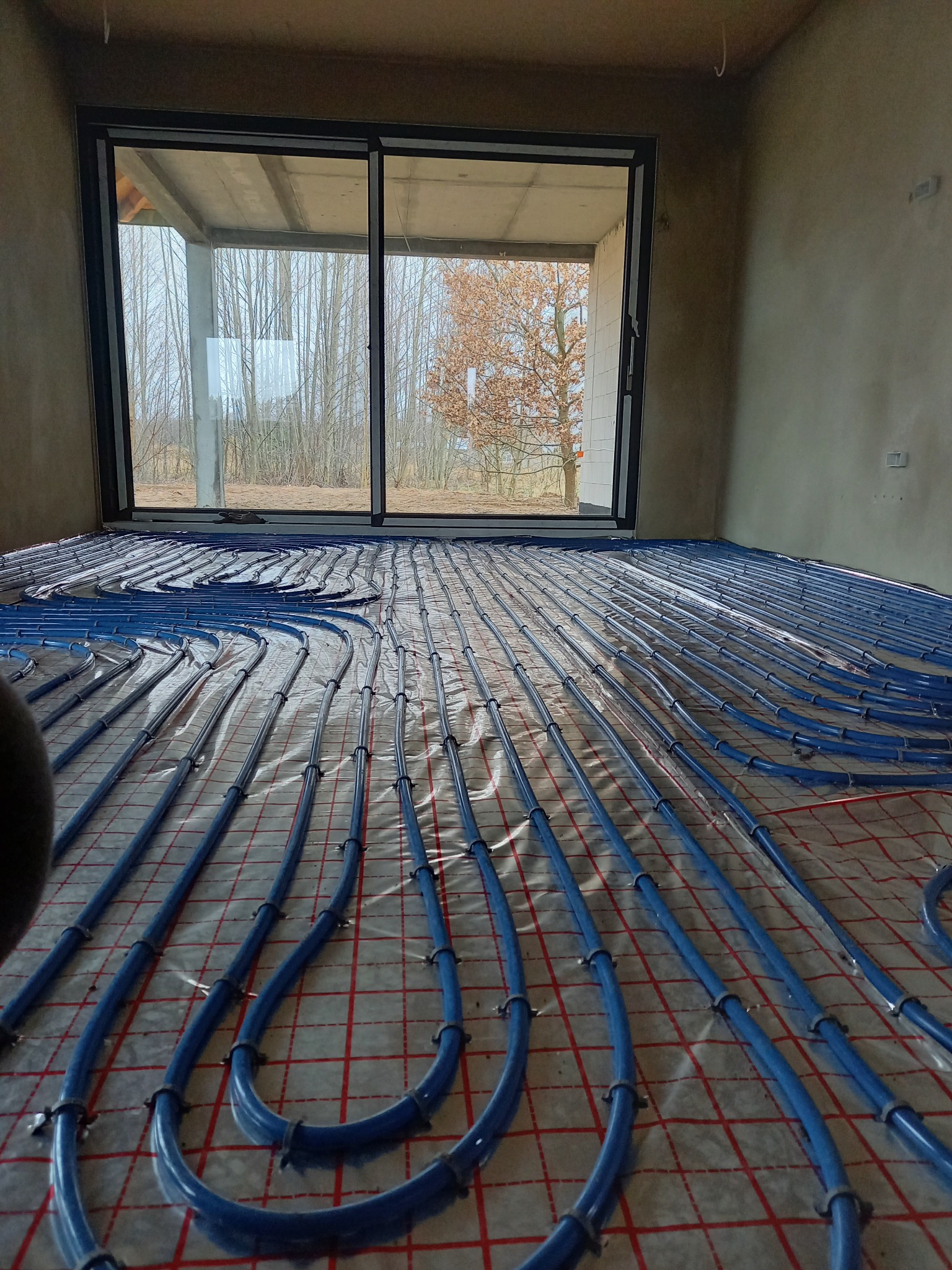 Niebieskie rury systemu grzewczego, zamocowane klamrami do podłoża z czerwoną kratką, rozłożone w surowym wnętrzu z dużym oknem na tle zimowego krajobrazu.