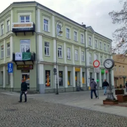 Ulica w mieście z budynkami o pastelowych elewacjach, brukowana nawierzchnia, zegar uliczny na słupie, widoczni przechodnie, pochmurne niebo.