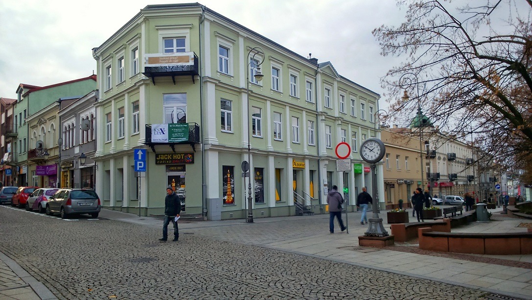 Ulica w mieście z budynkami o pastelowych elewacjach, brukowana nawierzchnia, zegar uliczny na słupie, widoczni przechodnie, pochmurne niebo.
