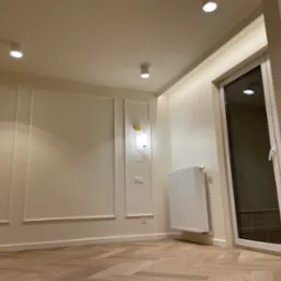 Jasne wnętrze po remoncie: parkiet w jodełkę, kremowe ściany z dekoracyjnymi listwami, nowoczesne oświetlenie sufitowe i ścienne, biały grzejnik oraz okno balkonowe.