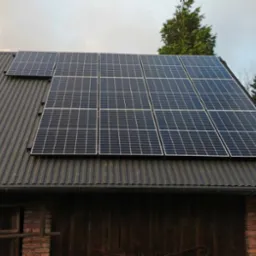 Panele słoneczne zamontowane na dachu z blachy trapezowej, widok na część elewacji budynku z cegły i drewniane wrota.