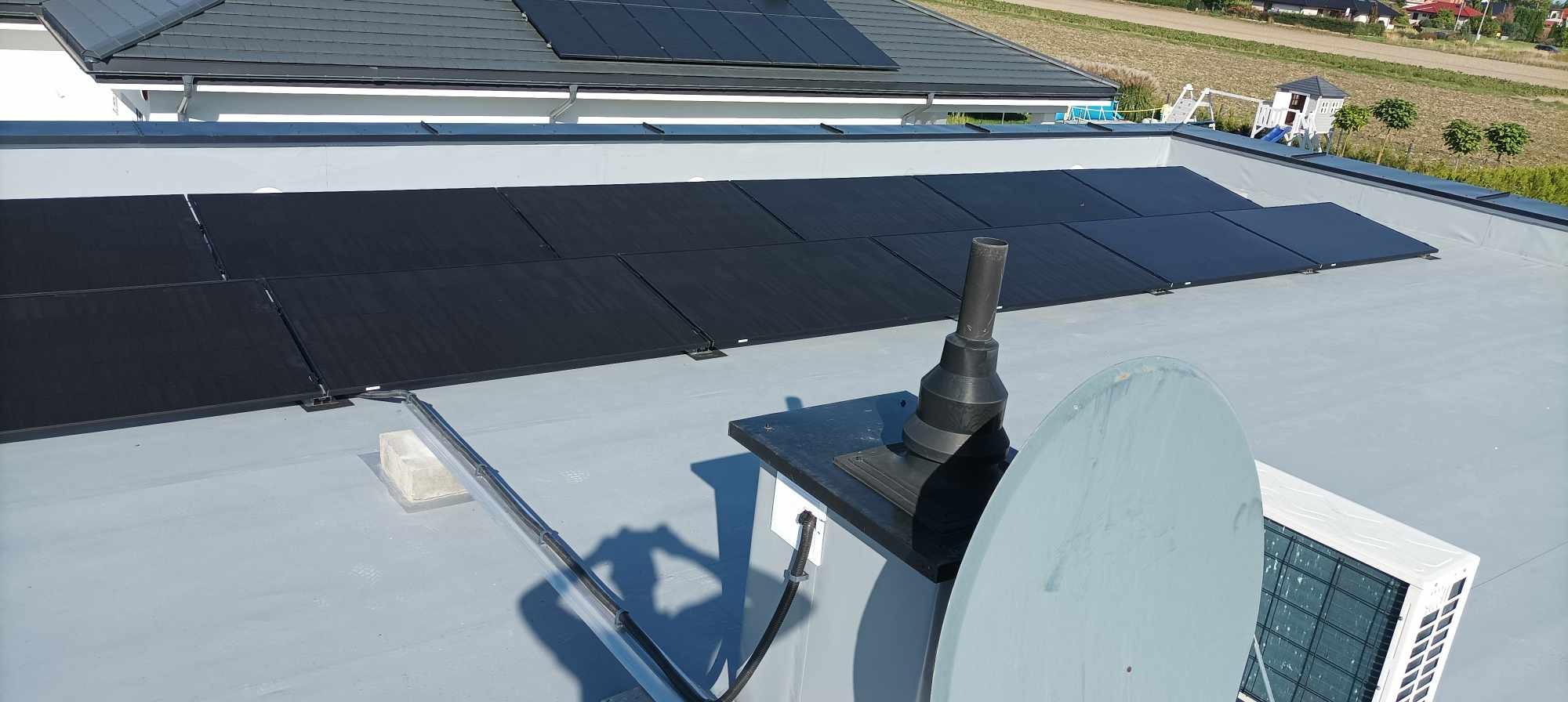 Panele słoneczne na płaskim dachu budynku jednorodzinnego, widoczna antena satelitarna i jednostka zewnętrzna klimatyzacji.