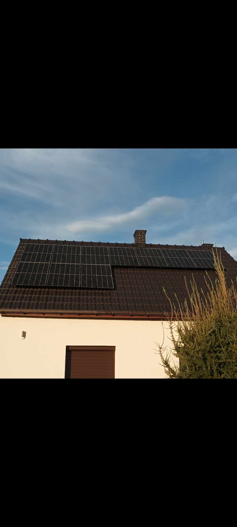 Panele fotowoltaiczne na brązowym dachu domu jednorodzinnego z jasną elewacją i brązową roletą okienną, widok z dołu w słoneczny dzień.