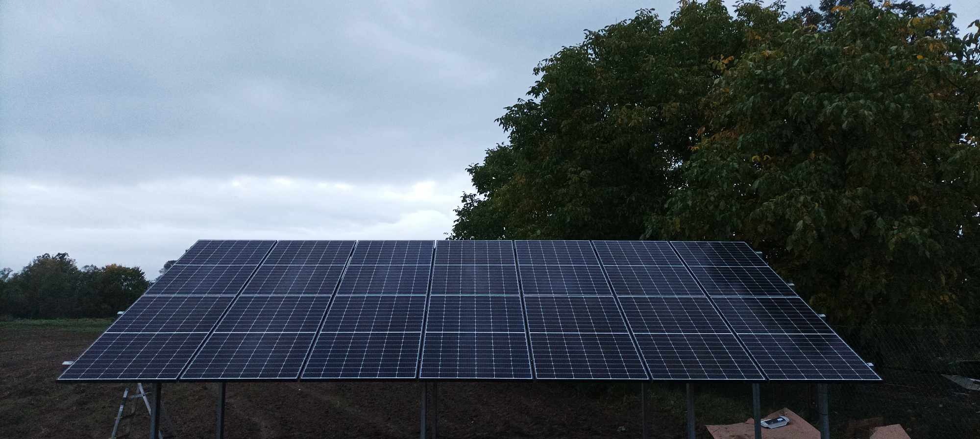 Zestaw paneli fotowoltaicznych na metalowej konstrukcji ustawiony na ziemi, w tle drzewo i pochmurne niebo.