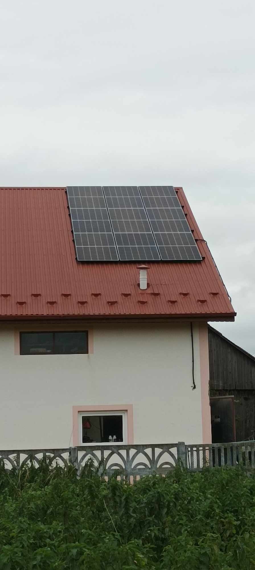 Panele słoneczne zamontowane na czerwonym, blaszanym dachu domu jednorodzinnego z kremową elewacją, widoczne krzewy w ogrodzie.