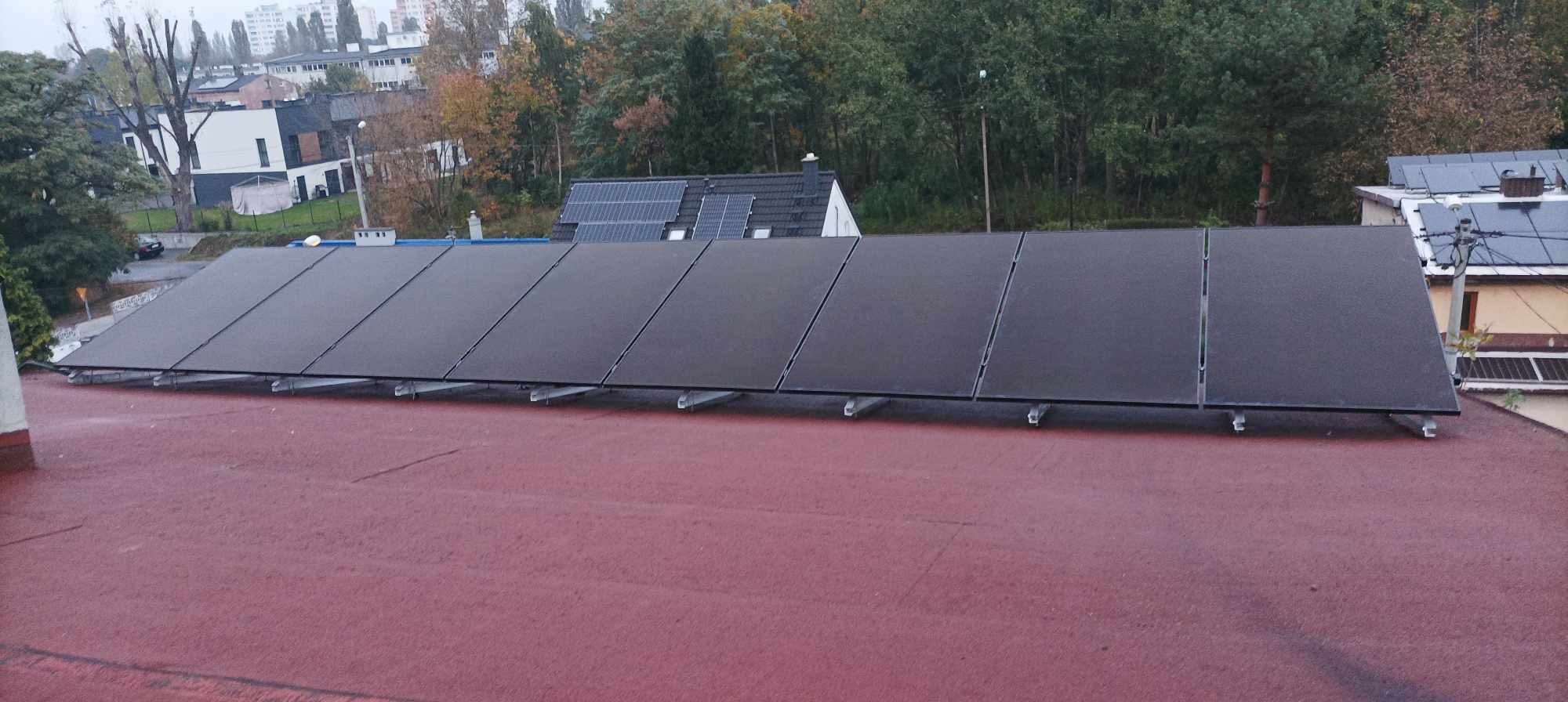Rząd paneli słonecznych zamontowanych na niskim dachu pokrytym czerwoną papą, z budynkami mieszkalnymi i drzewami w tle.