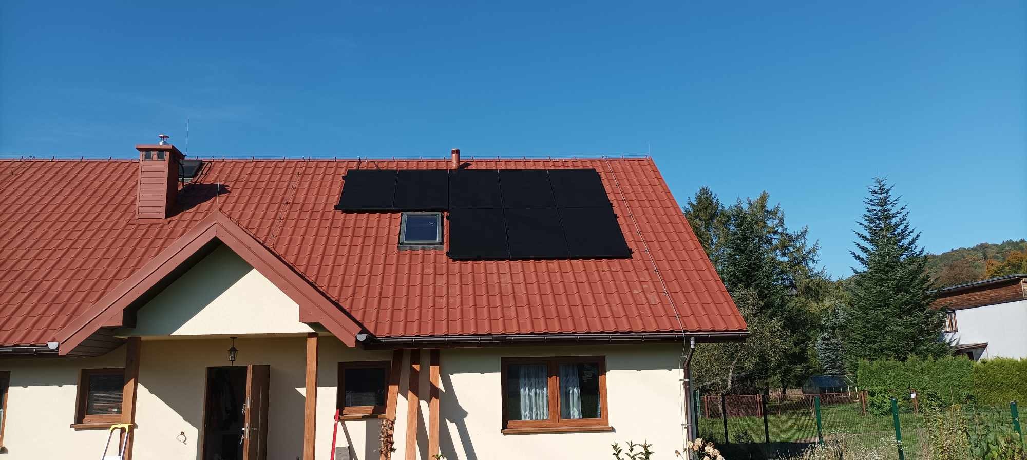 Instalacja fotowoltaiczna na dachu domu jednorodzinnego z czerwoną dachówką, widoczne panele słoneczne i okno dachowe, błękitne niebo w tle.