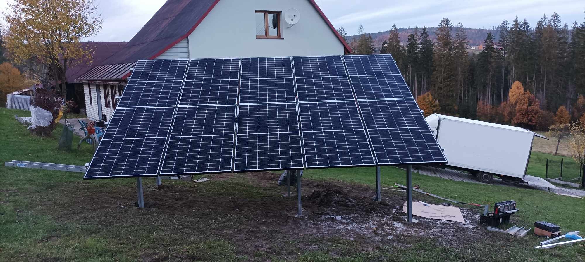 Panel fotowoltaiczny zainstalowany na metalowych słupkach na tle domu jednorodzinnego i lasu, widoczny także samochód dostawczy i drabina.