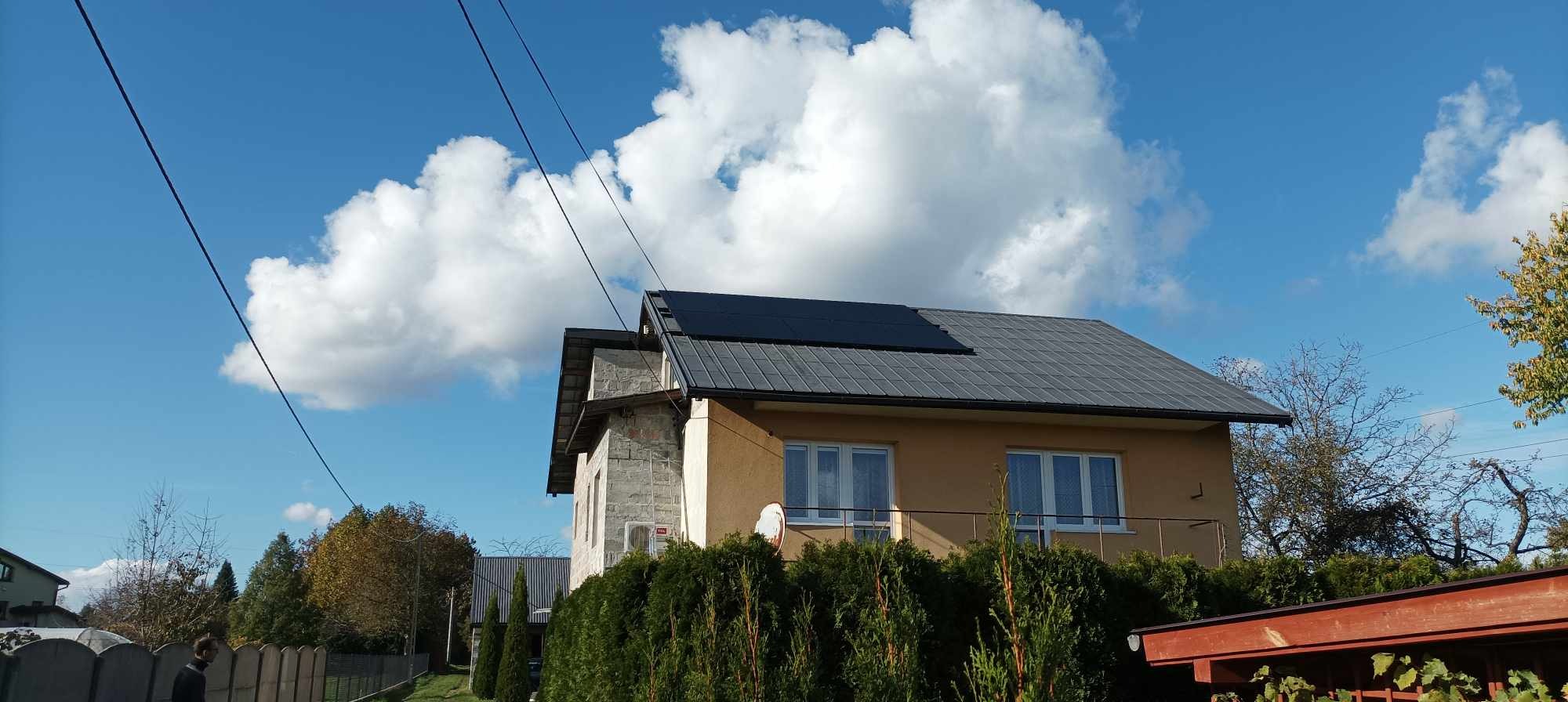 Dom jednorodzinny z panelami słonecznymi na szarym dachu pod błękitnym niebem z białymi chmurami, widoczne również przewody elektryczne i iglaki w ogrodzie.