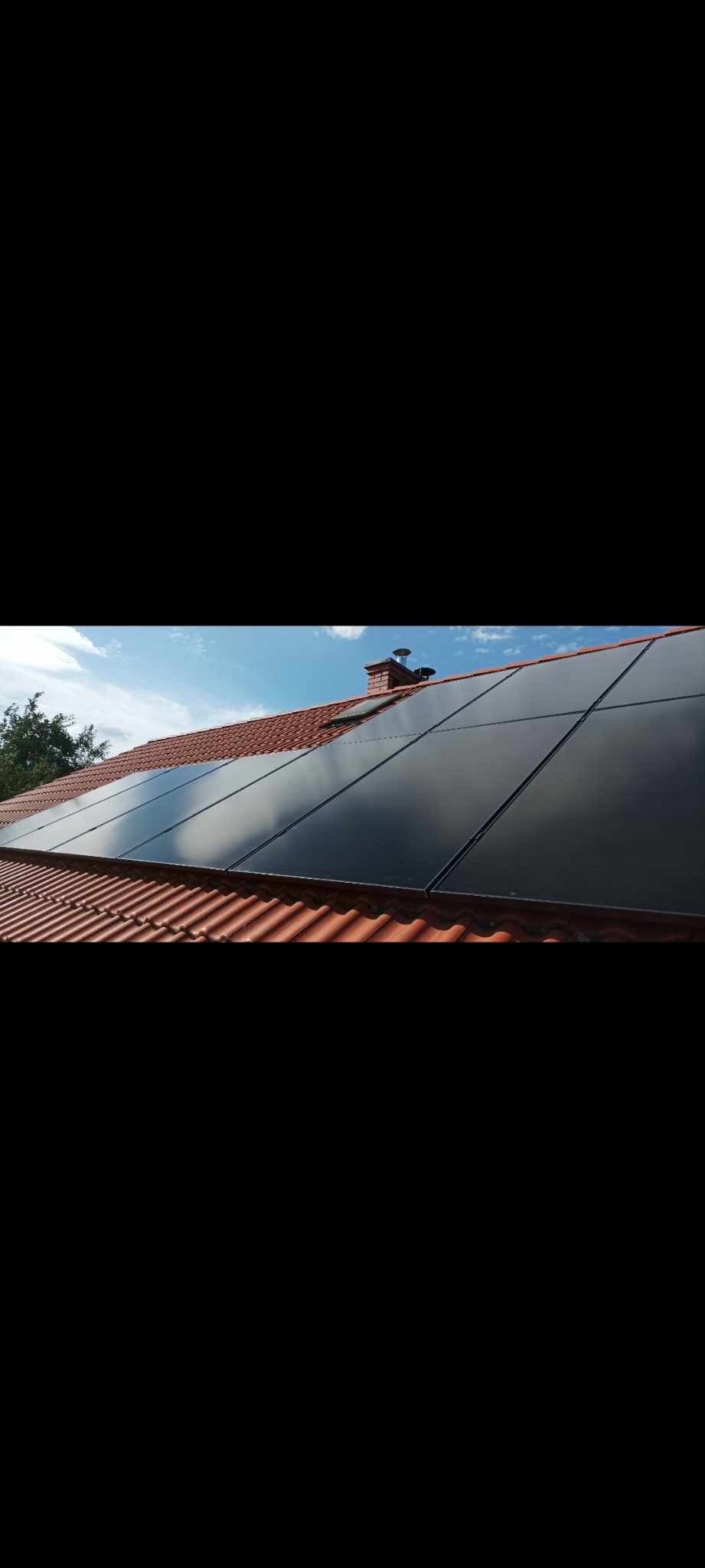 Czarne panele słoneczne na spadzistym dachu z czerwonej dachówki, widoczne drzewo i błękitne niebo z chmurami w tle.