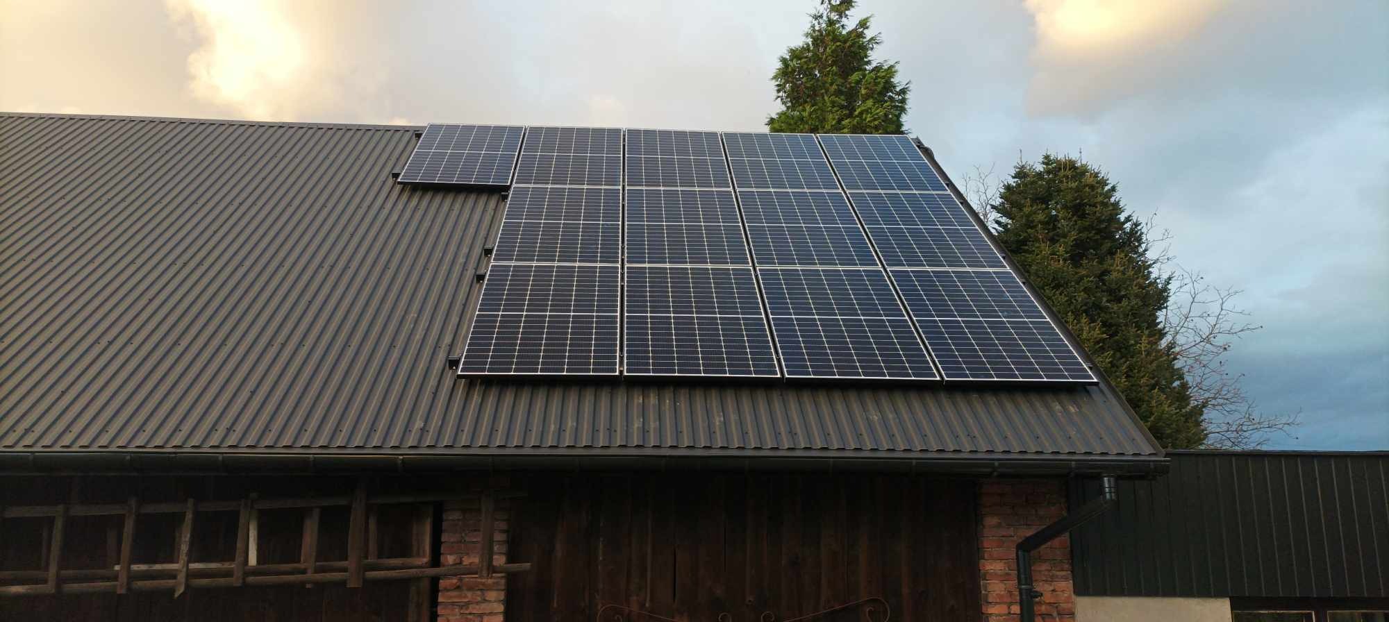 Panele słoneczne zamontowane na dachu z blachy trapezowej, widok na część elewacji budynku z cegły i drewniane wrota.