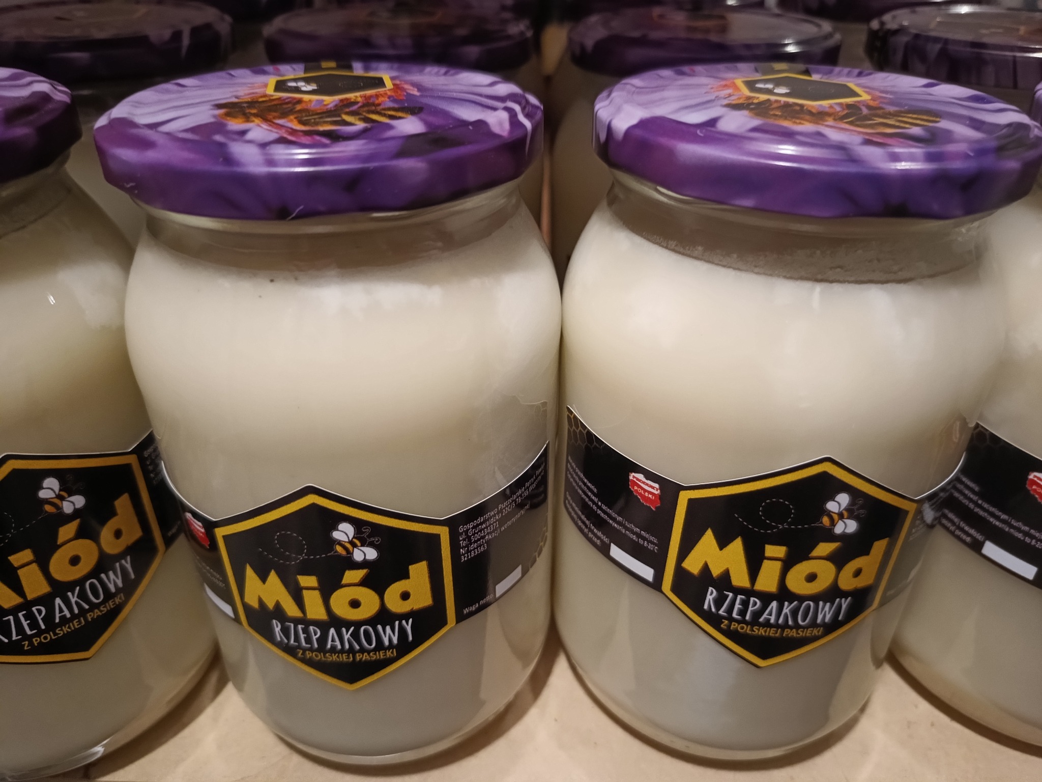 Słoiki z miodem rzepakowym, etykiety z motywem pszczoły, fioletowe wieczka z kwiatowym wzorem, ustawione rzędem na półce.