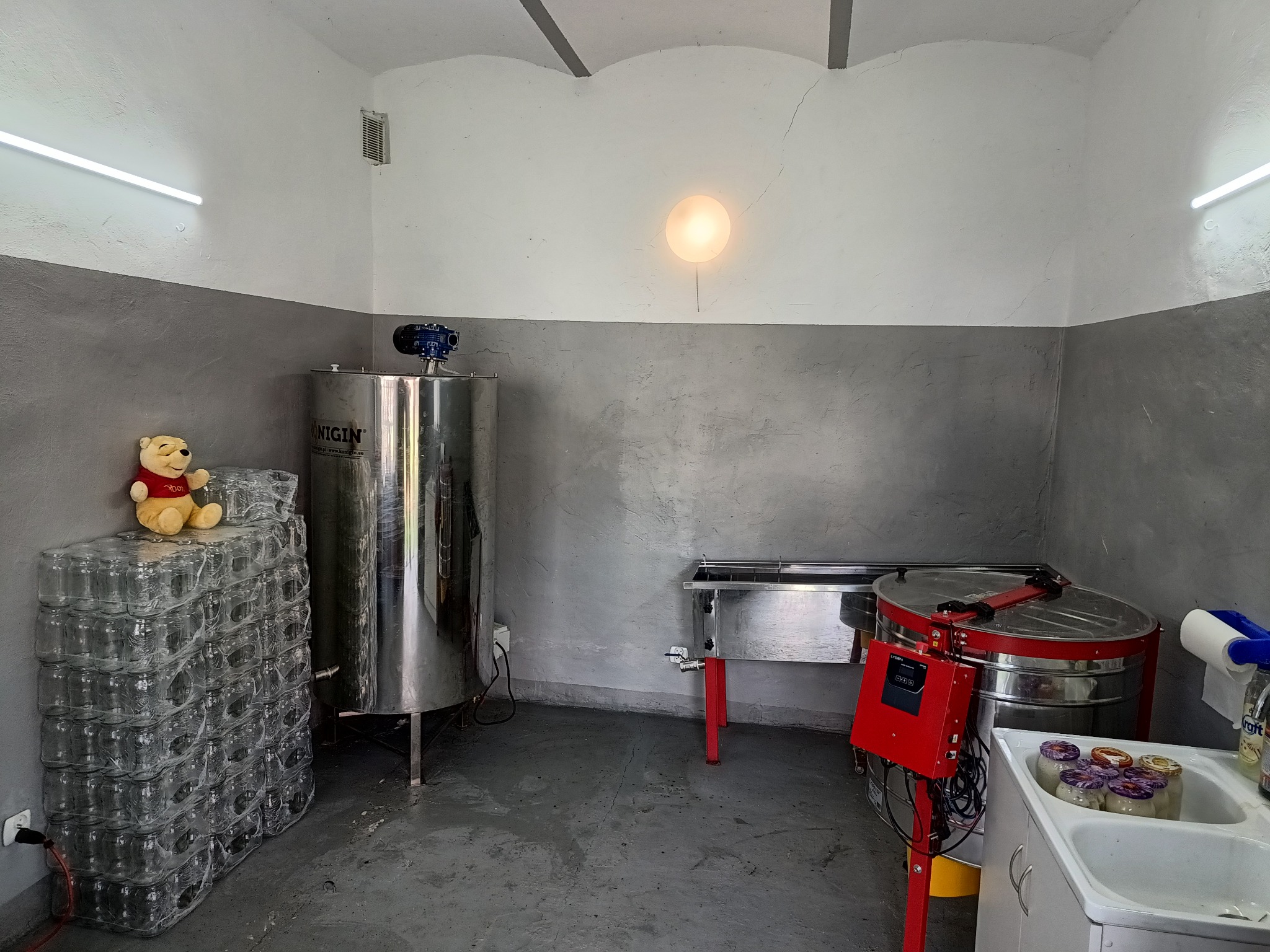 Wnętrze pracowni z urządzeniami do produkcji, z bliska widoczne zapakowane słoiki na miod, stalowy zbiornik i miodarka z czerwonym panelem sterowania, a na słoikach siedzi pluszowy Kubuś Puchatek.