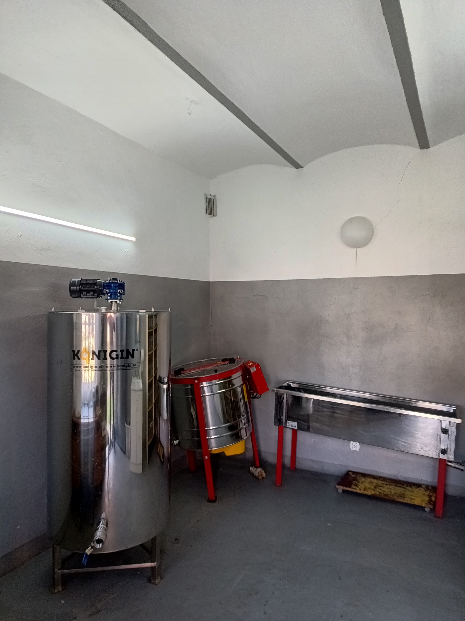 Wnętrze pracowni pszczelarskiej z nierdzewnymi urządzeniami: odstojnik z silnikiem, miodarka i stół do odsklepiania plastrów na czerwonych nogach.