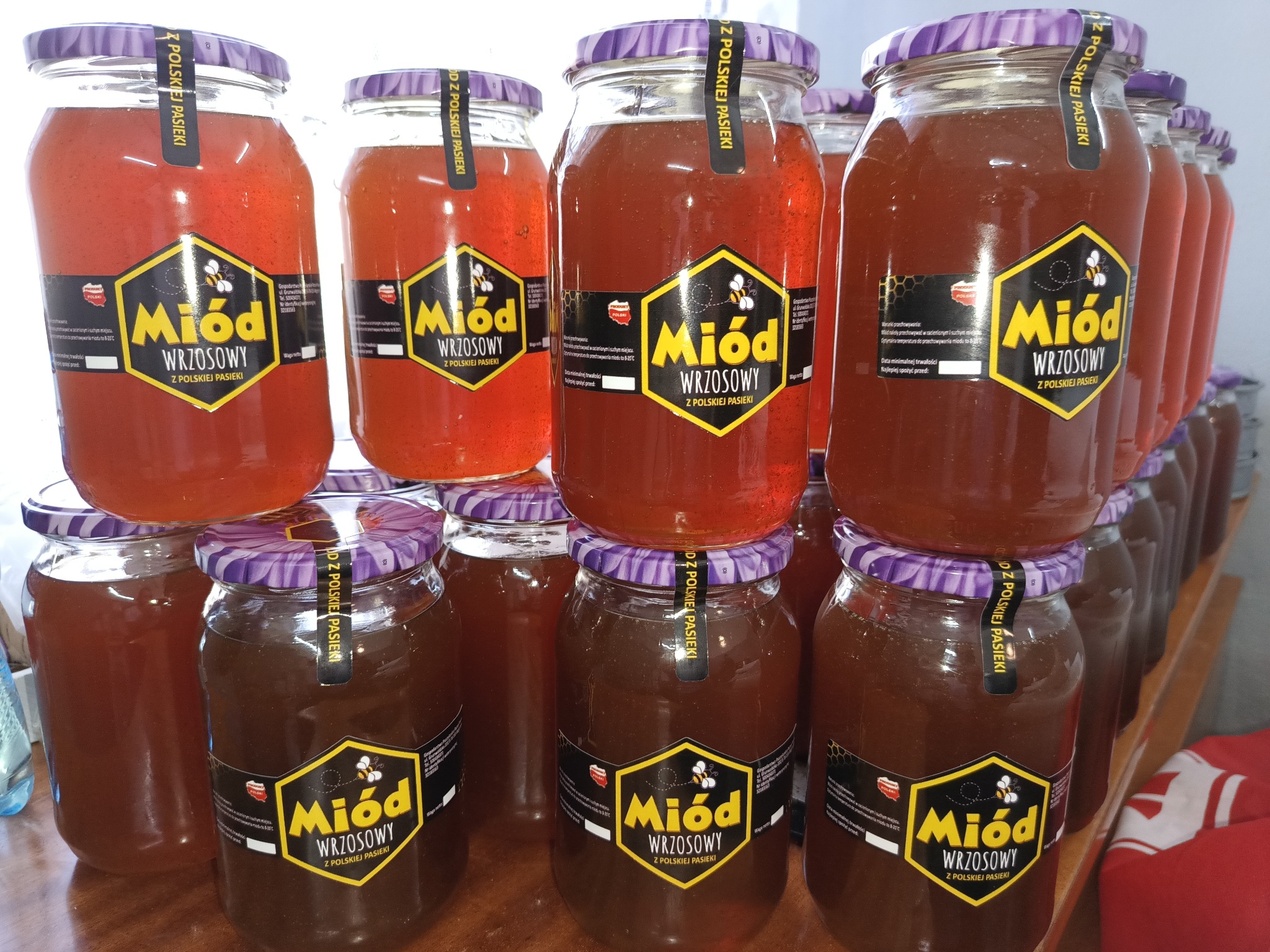 Słoiki z miodem wrzosowym ustawione w rzędach, widoczne etykiety z nazwą i logo producenta, różne odcienie miodu w słoikach.