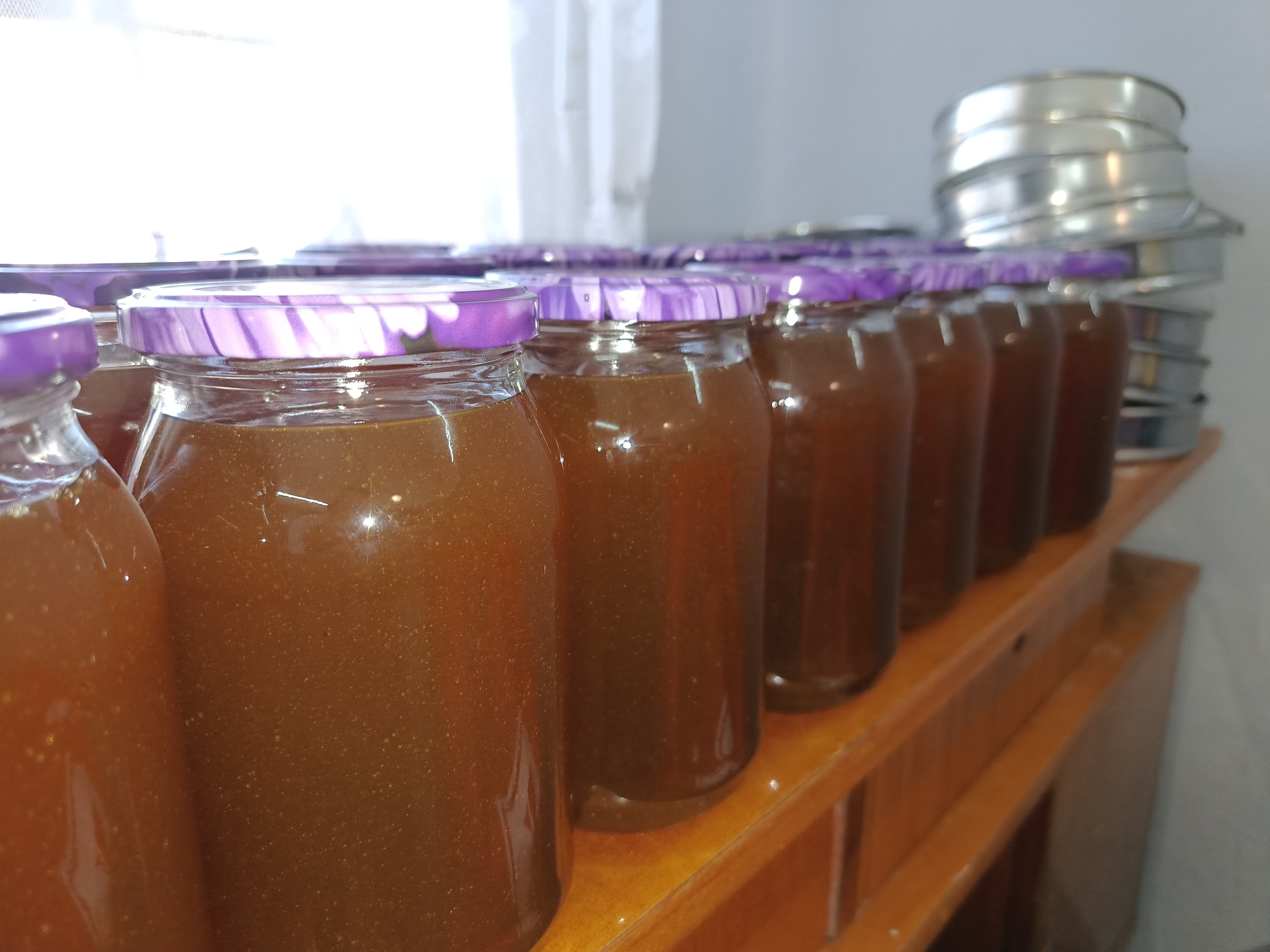 Rząd słoików z ciemnym płynem, zamkniętych fioletowymi wieczkami, ustawionych na drewnianej półce, w tle stos metalowych naczyń.