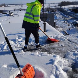 3Miasto-Hol Piotr Szpreniec - Pracownik w odblaskowej kurtce odśnieża płaski dach budynku za pomocą pomarańczowej łopaty. W tle widoczny krajobraz miejski pod błękitnym niebem.