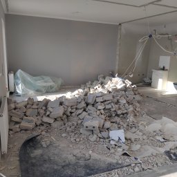 3Miasto-Hol Piotr Szpreniec - Wnętrze po wyburzeniu ściany: sterta gruzu z pustaków na podłodze, widoczne instalacje elektryczne i fragmenty ścian. Remont w trakcie, surowe wykończenie.