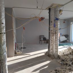 3Miasto-Hol - Wnętrze w trakcie demontażu: odsłonięte przewody, zabezpieczone rury, gruz. Widoczne narzędzia i fragmenty ścian. Jasne światło podkreśla surowość stanu obecnego.